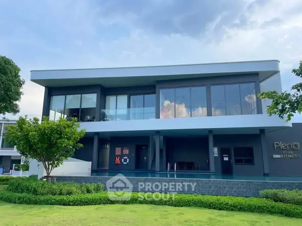 Modern fitness club building with large glass windows and lush green surroundings.