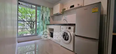 Modern kitchen with washing machine and fridge, large window with leafy view.