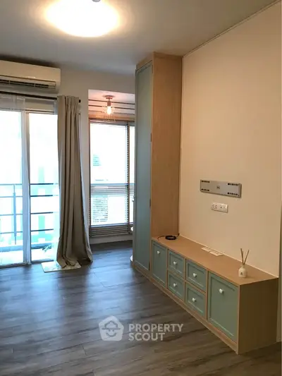 Modern living room with wooden flooring and balcony access, featuring built-in storage and air conditioning.