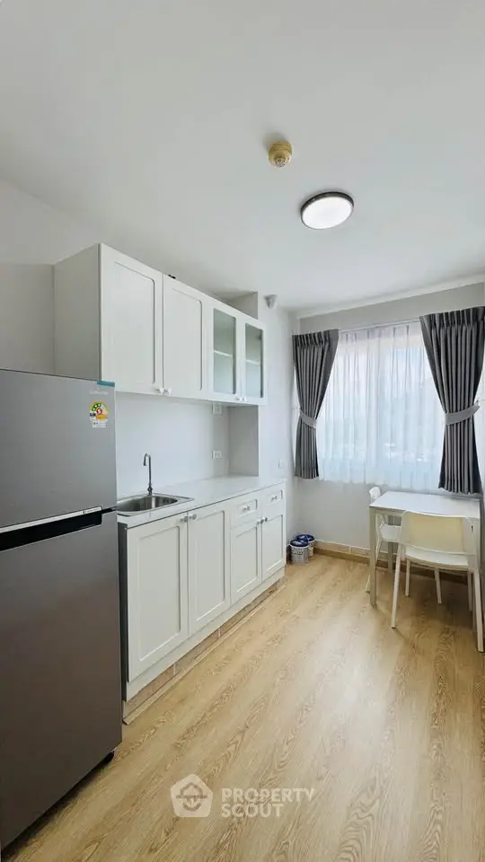 Modern kitchen with sleek white cabinets and natural light, featuring a compact dining area and stainless steel fridge.