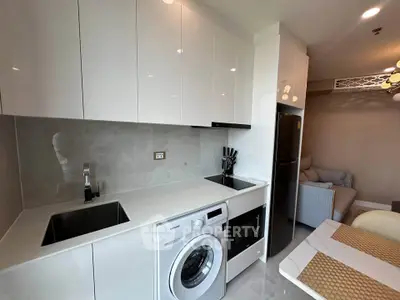 Modern kitchen with sleek white cabinets, washing machine, and cozy living area in open layout apartment.