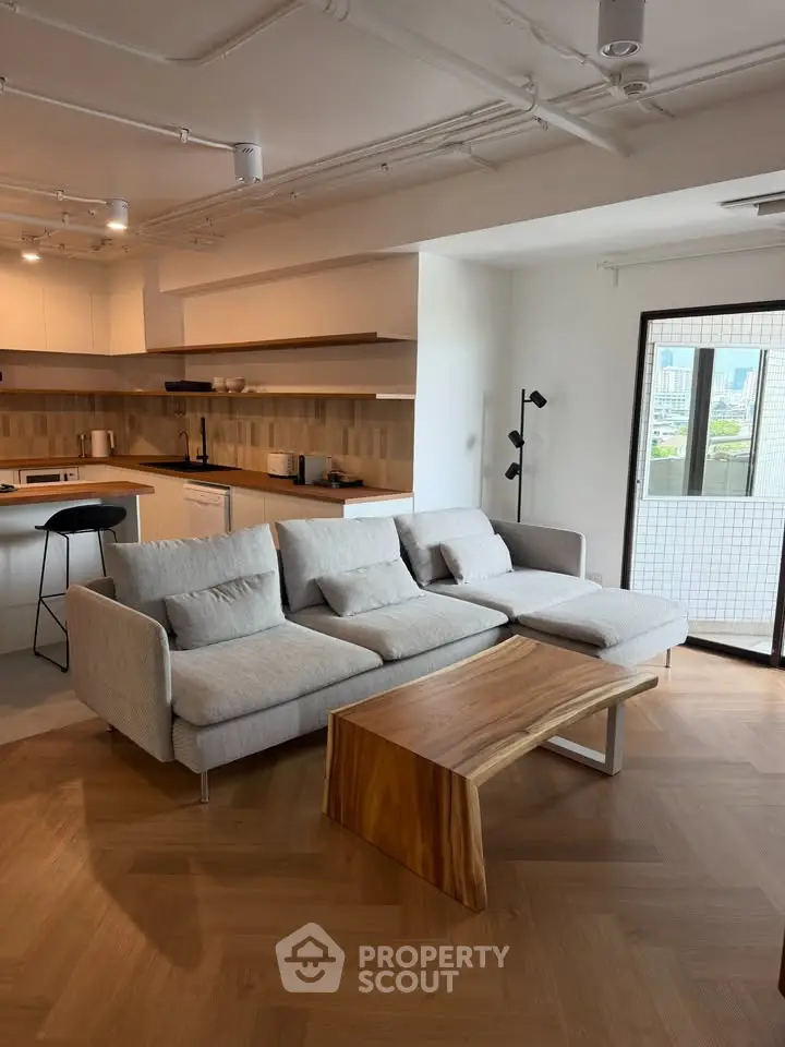 Modern living room with open kitchen and city view balcony, featuring sleek furniture and wooden flooring.