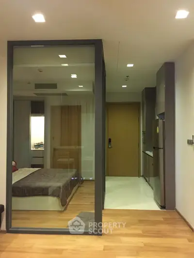 Modern studio apartment with glass partition, wooden flooring, and compact kitchen area.