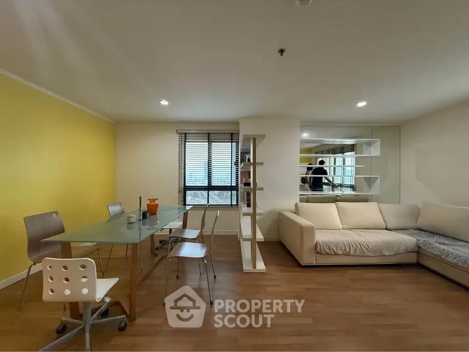 Modern living room with glass dining table and cozy sofa, featuring a bright yellow accent wall.