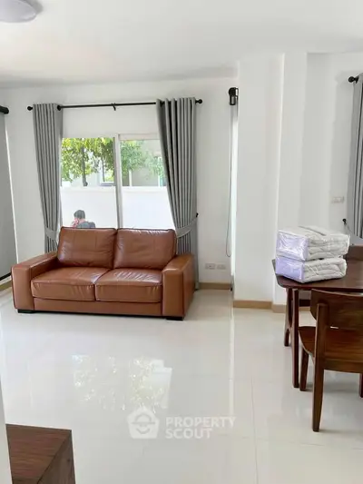 Bright living room with brown leather sofa and dining table, featuring large window and elegant curtains.