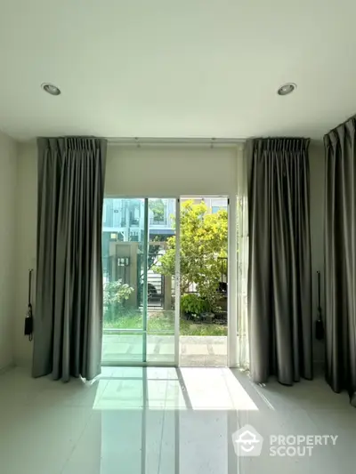 Bright room with large sliding glass doors opening to a lush garden view, featuring elegant curtains and glossy flooring.