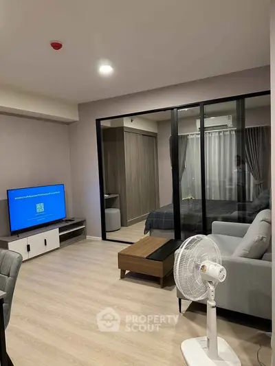Modern living room with glass partition to bedroom, featuring sleek furniture and light wood flooring.