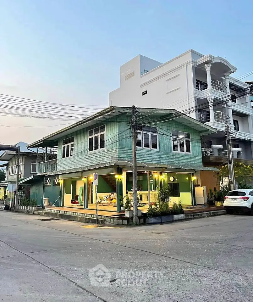 Charming two-story green wooden house on a corner lot with modern white building backdrop.