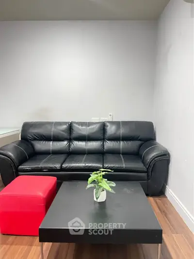 Modern living room with black leather sofa and red ottoman on wooden floor.