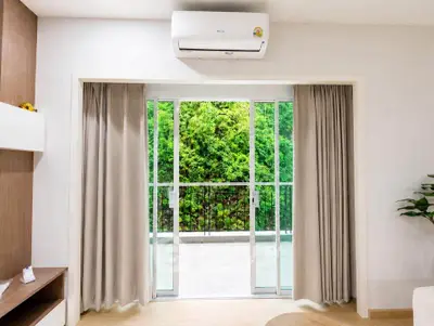 Modern living room with sliding glass doors opening to lush greenery, featuring air conditioning and elegant curtains.