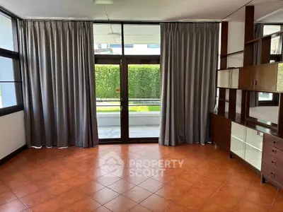 Spacious living room with large windows and elegant curtains, featuring built-in wooden shelves and terracotta tile flooring.