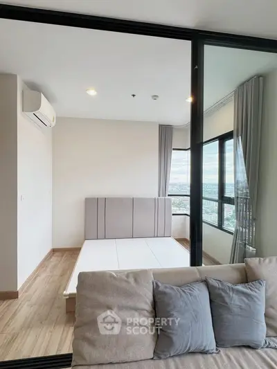 Modern bedroom with large windows and city view, featuring a minimalist bed frame and cozy sofa.