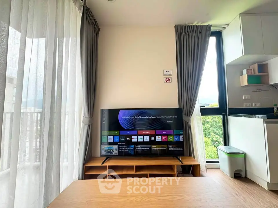 Modern living room with large TV, elegant curtains, and partial kitchen view in a bright apartment.