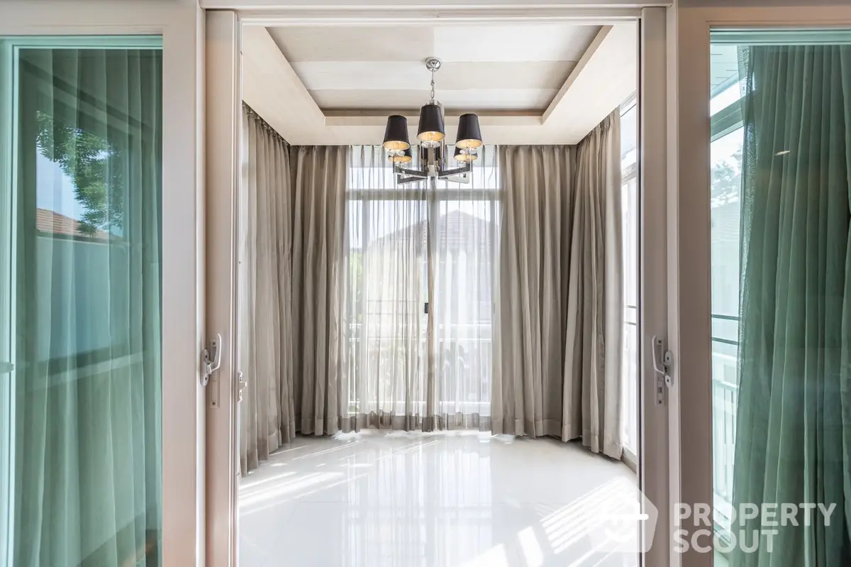 Sunlit spacious room with gleaming floor tiles, elegant chandelier, and floor-to-ceiling drapes opening to a serene balcony view.