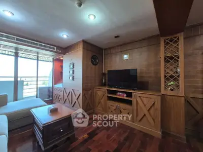 Spacious living room with wooden accents and large TV, opening to a balcony with natural light.
