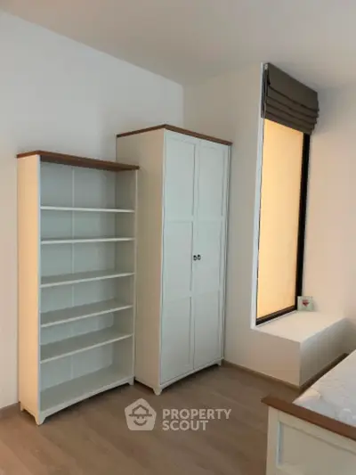 Bright and cozy bedroom with ample storage, featuring a large wardrobe and a bookshelf, complemented by warm wooden flooring and a large window for natural light.