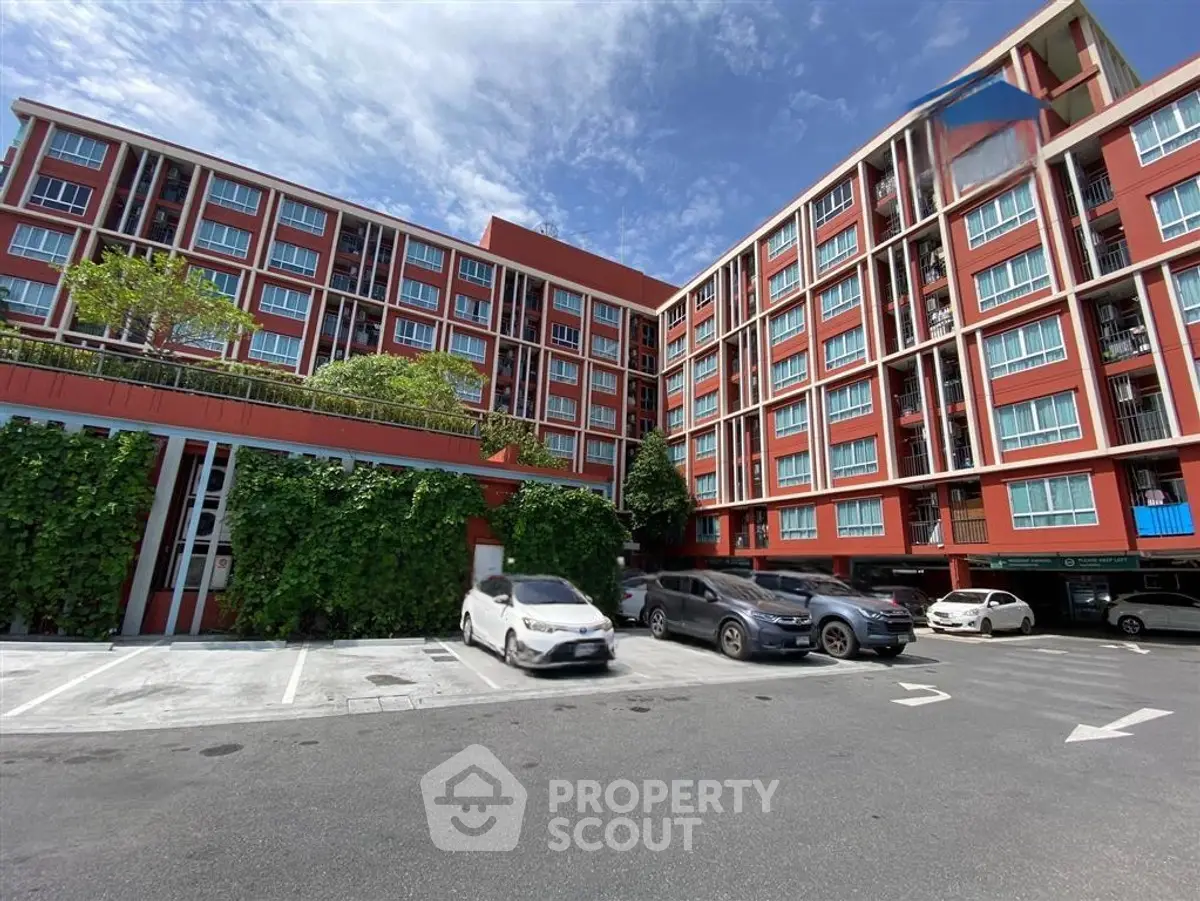 Modern red apartment building with lush greenery and spacious parking area under a clear blue sky.