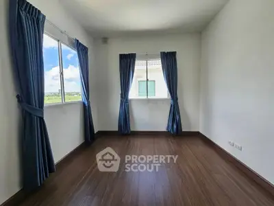 Spacious empty room with wooden flooring and large windows with blue curtains.