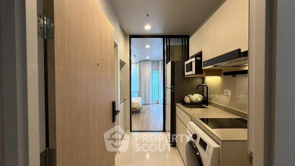 Modern apartment entrance with sleek kitchen and washing machine, leading to a cozy bedroom.