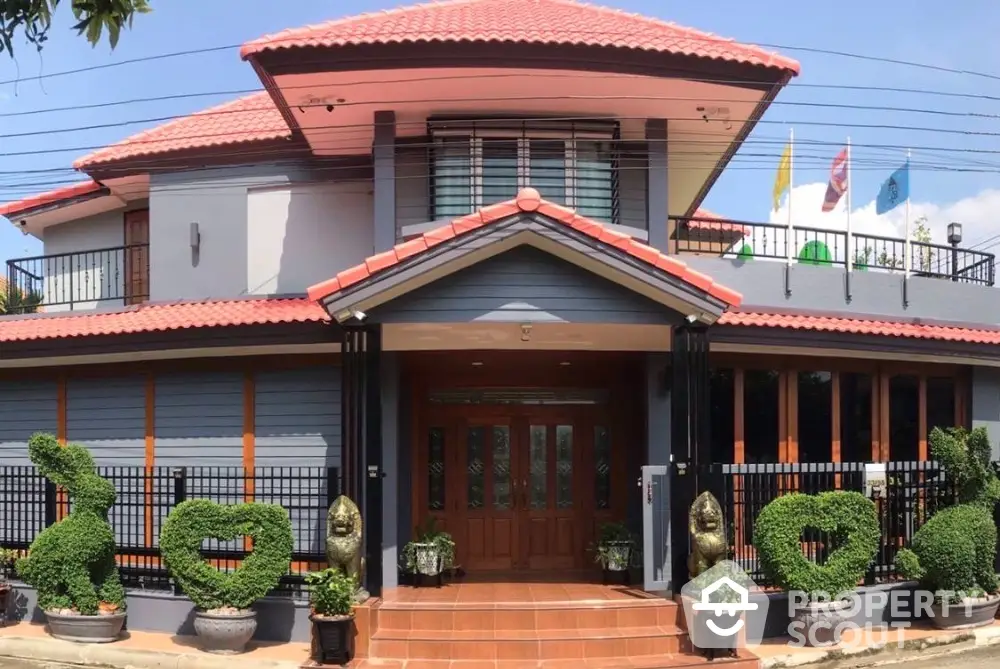 Charming two-story house with red-tiled roof and manicured garden, featuring heart-shaped topiary and elegant entrance.