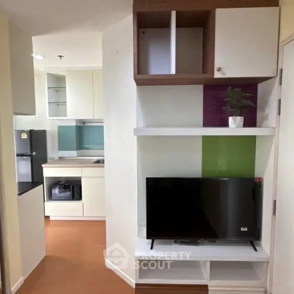 Modern apartment interior with compact kitchen and living area featuring sleek cabinetry and a flat-screen TV.