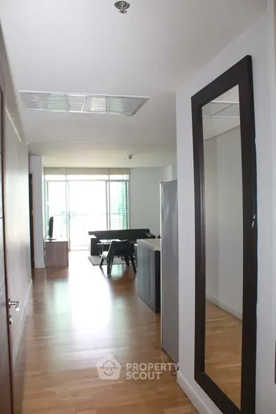 Modern apartment interior with wooden flooring and large mirror, leading to a bright living area with balcony access.