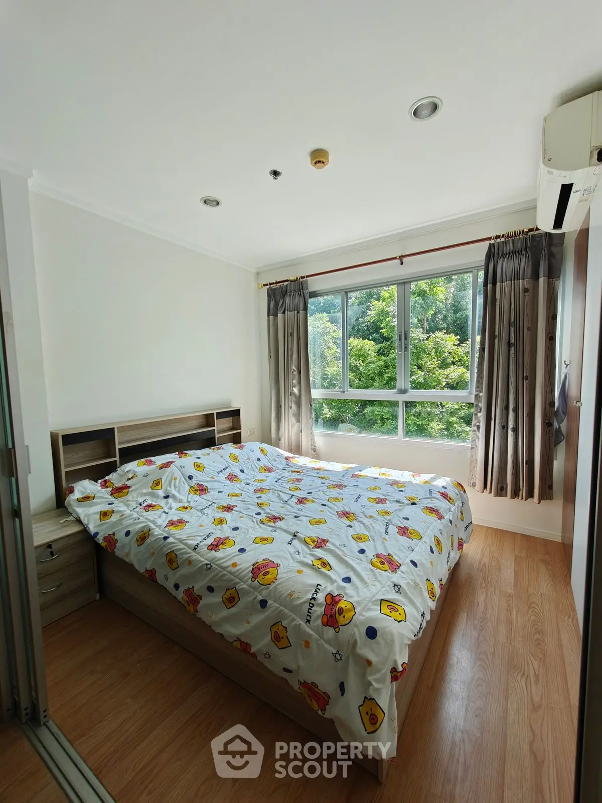 Bright bedroom with large window and wooden flooring, featuring a cozy bed with colorful bedding.