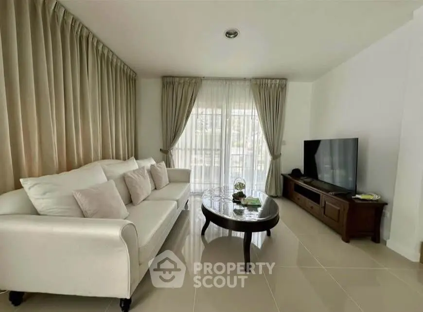 Elegant living room with white sofa, large TV, and natural light from floor-to-ceiling windows.