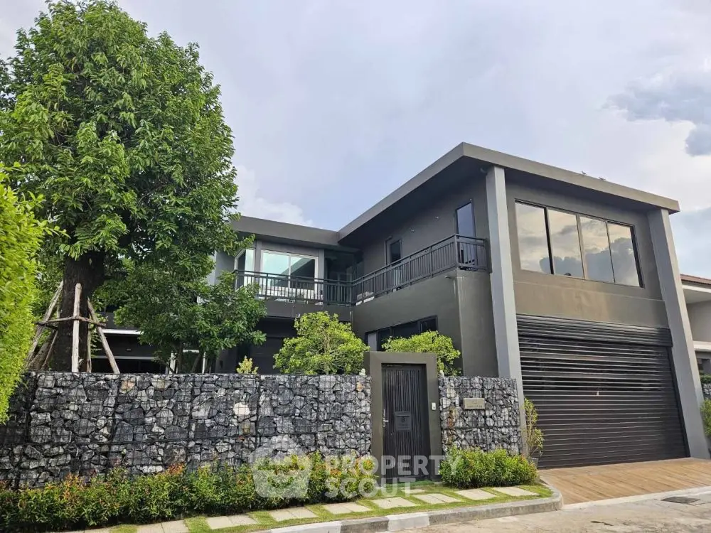 Modern two-story house with large windows and stone fence, surrounded by lush greenery.