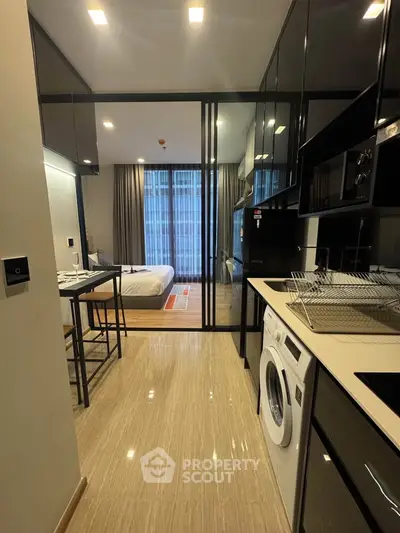 Modern studio apartment with sleek kitchen and cozy bedroom, featuring glass partition and washing machine.
