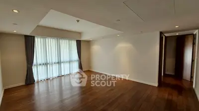 Spacious empty room with wooden flooring and large window with curtains.