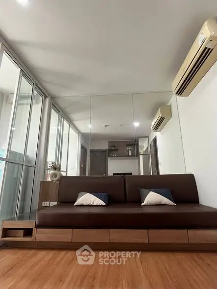 Modern living room with sleek brown sofa and mirrored wall, featuring air conditioning and wooden flooring.