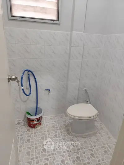 Compact bathroom with tiled walls and floor, featuring a toilet and cleaning supplies.