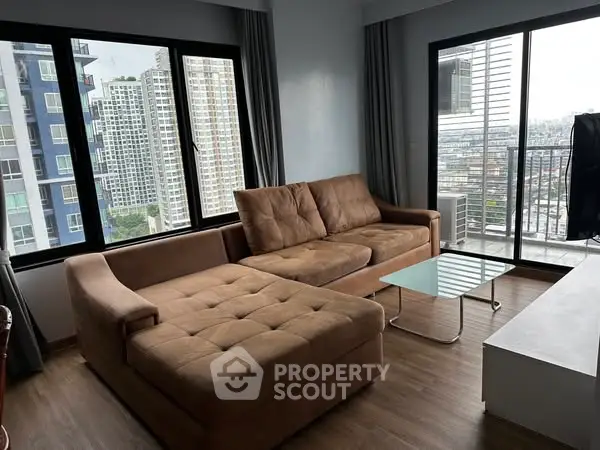 Modern living room with large windows and city view, featuring a cozy brown sectional sofa and glass coffee table.