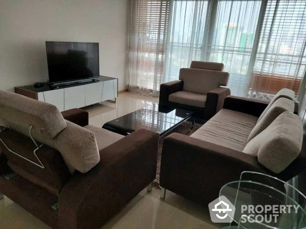 Spacious and well-lit living room with comfortable brown sofas, glass coffee table, and a modern television set, perfect for family relaxation.