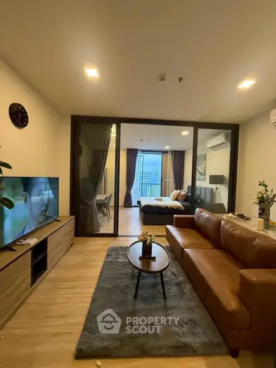 Spacious modern living room with leather sofa and TV, leading to a cozy bedroom with city view.