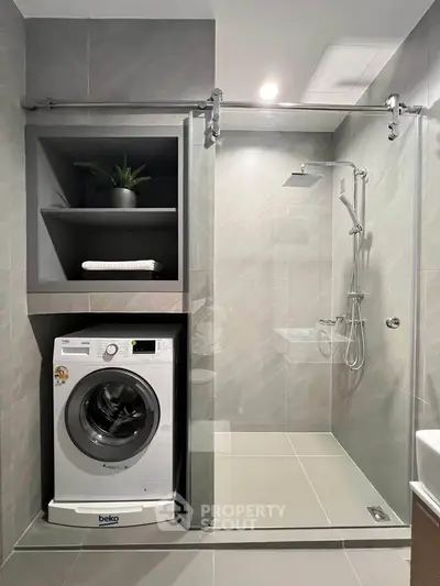Modern bathroom with sleek shower and built-in washing machine, featuring elegant gray tiles and recessed shelving.