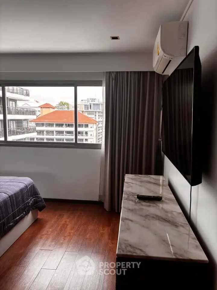 Modern bedroom with large window and city view, featuring a wall-mounted TV and air conditioning.