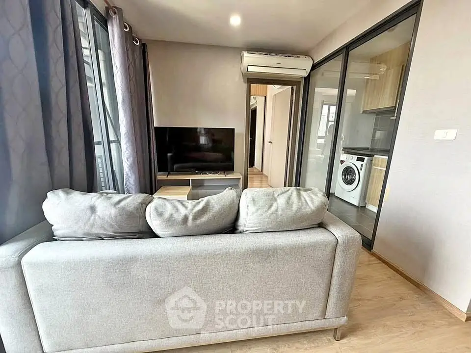 Modern living room with gray sofa, TV, and view into kitchen with washing machine.