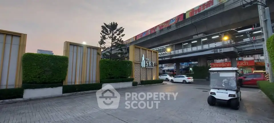 Modern urban entrance with lush greenery and elevated train tracks nearby.