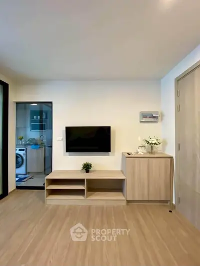 Modern living room with wooden flooring and TV, adjacent to a kitchen with washing machine.