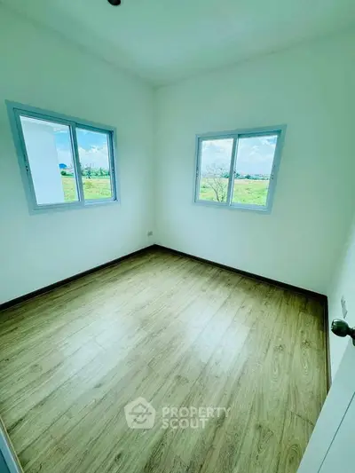 Bright empty room with wooden flooring and two windows offering scenic views.