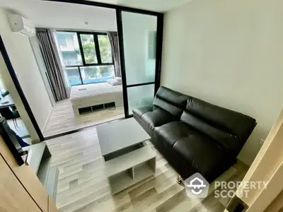 Modern living room with sleek black leather sofa and minimalist coffee table, leading to a cozy bedroom with natural light.