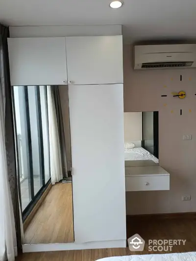 Modern bedroom with sleek wardrobe and air conditioning unit, featuring balcony access and wooden flooring.