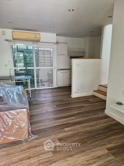 Spacious living room with modern flooring and natural light, featuring a compact kitchen and staircase.