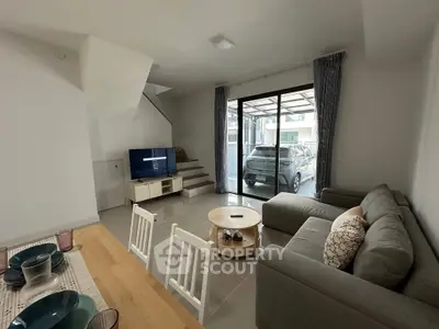Modern living room with cozy sofa, TV, and dining area, featuring large windows and a view of the carport.