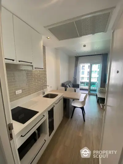 Modern kitchen with sleek white cabinetry and built-in appliances seamlessly opens to a cozy living area, leading to a sunlit balcony.