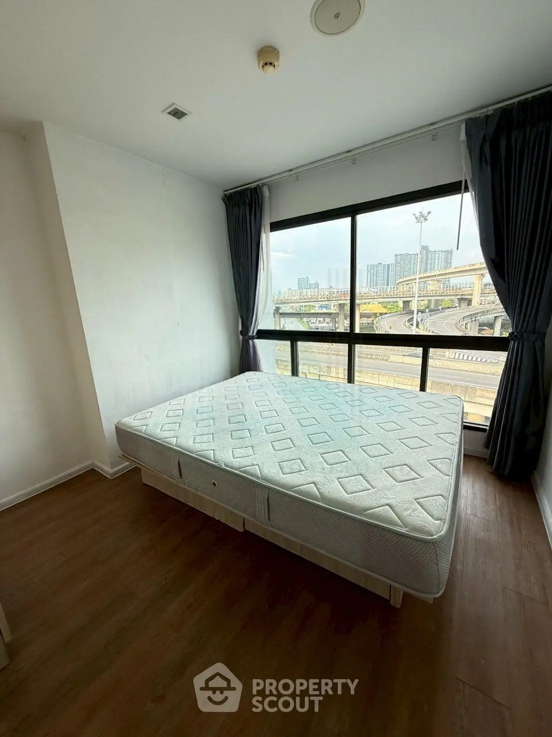 Spacious bedroom with large window offering city view, featuring a comfortable mattress and elegant wooden flooring.