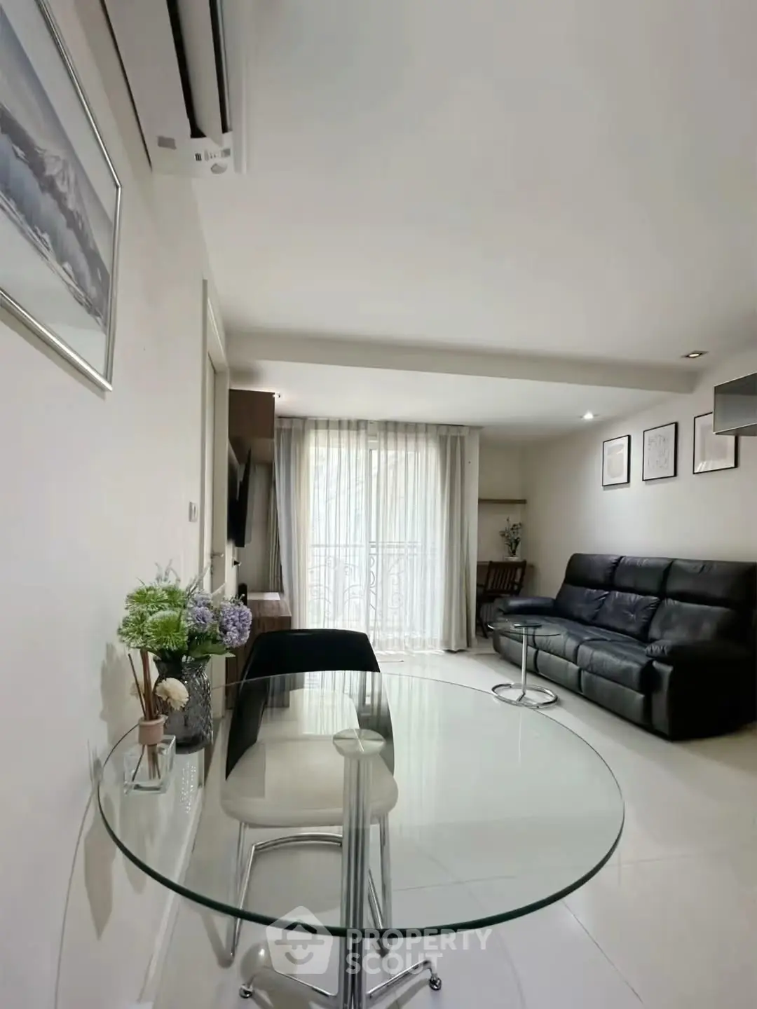 Modern living room with glass table, black leather sofa, and natural light from large window.
