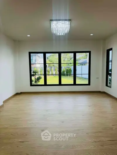 Spacious empty room with large windows overlooking a lush garden, featuring elegant wooden flooring.